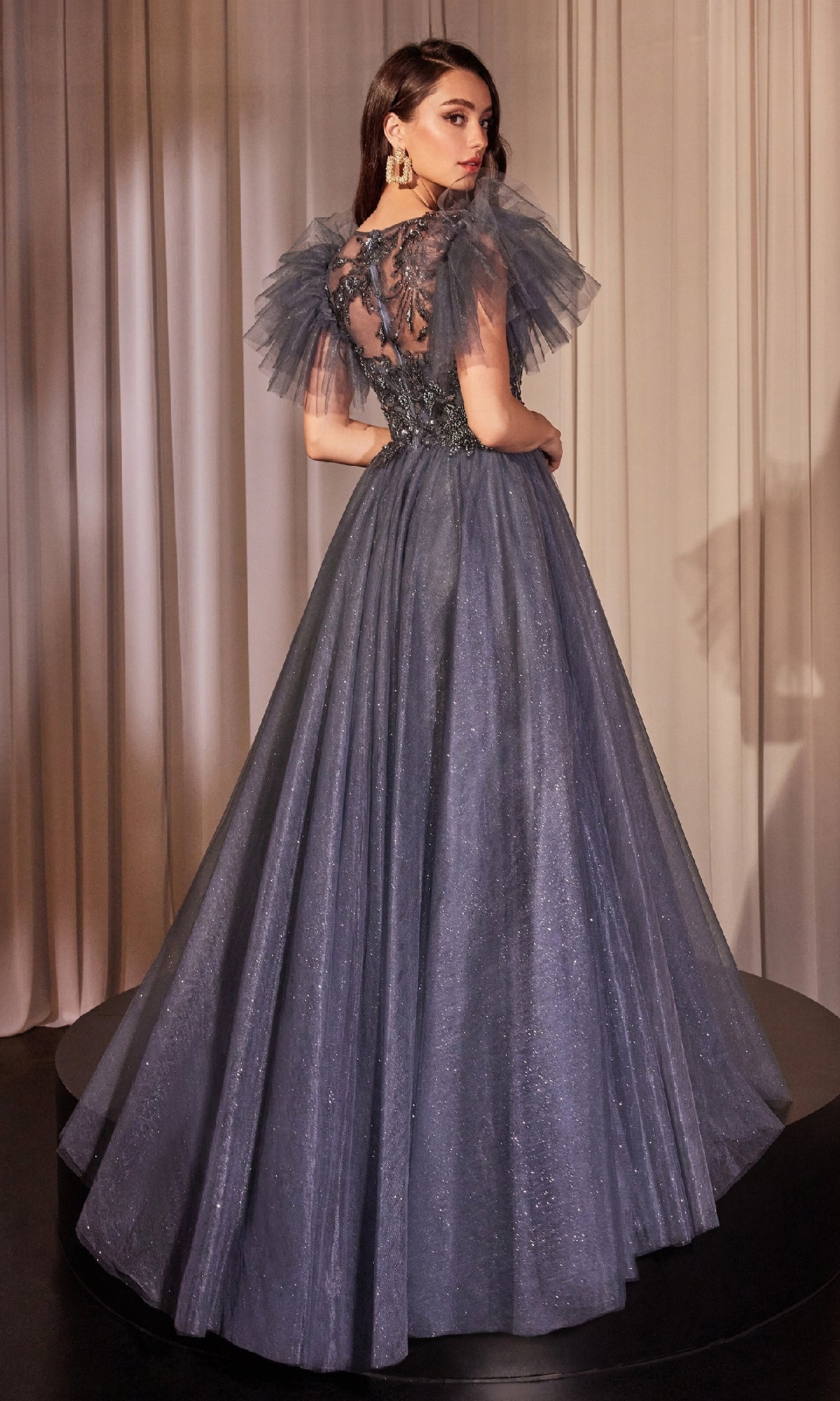 A woman poses in front of cream curtains wearing the Ladivine CM359 Flutter-Sleeve Blue Prom Ball Gown, a floor-length dress featuring shimmery fabric, graceful flutter sleeves, and intricate back detailing.